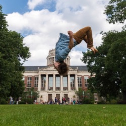 A person is mid-air performing a backflip on a grassy field with a large, stately building with columns and a dome in the background. 的 sky is partly cloudy and there are several people and trees in the vicinity.